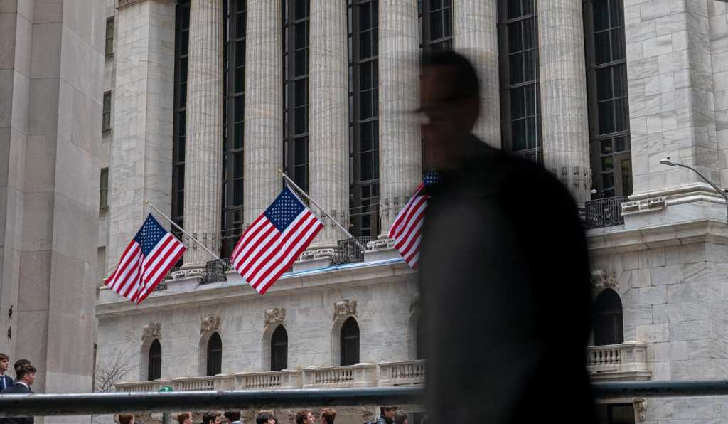 A large classical building with tall, fluted columns and arched windows, adorned with three American flags hanging on poles. In the foreground, a blurred silhouette of a person walking obscures part of the view. Several people are visible near the bottom of the image, standing or walking along the street.