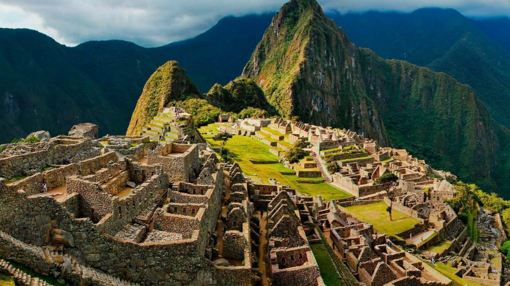 Ancient stone ruins of Machu Picchu, an Incan citadel, set against steep green mountains under a cloudy sky. The site features terraced fields, stone buildings, and pathways.