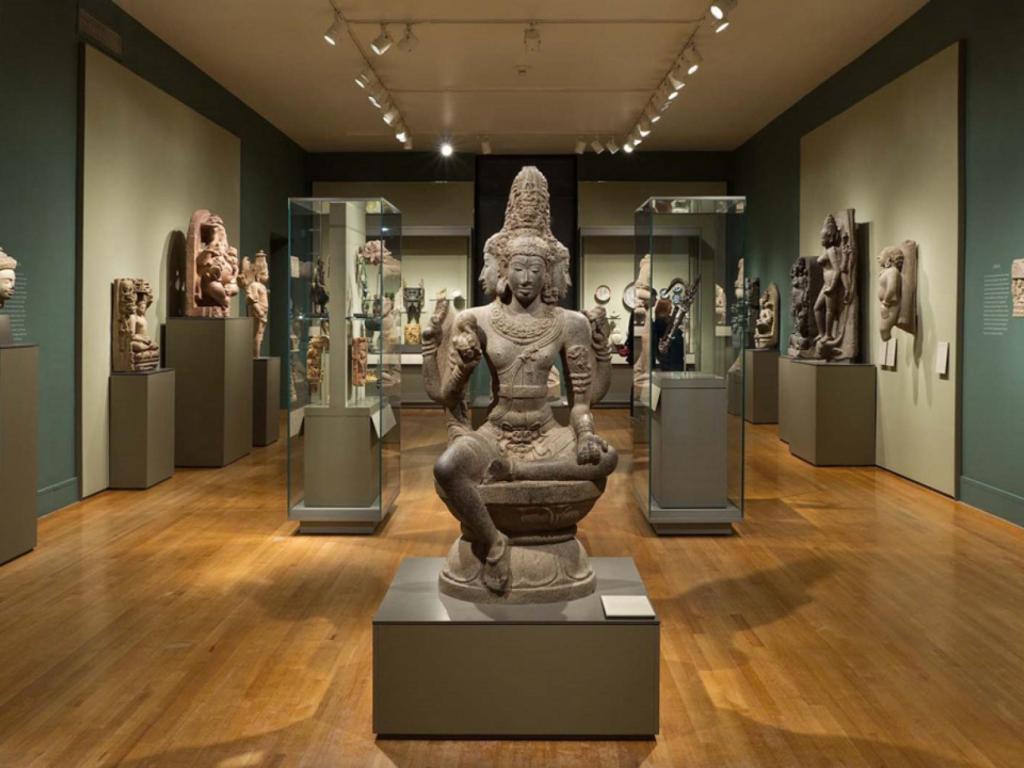 A large stone sculpture of a multi-armed deity seated cross-legged on a pedestal is displayed prominently in the center of a museum gallery. The deity has an elaborate headdress and detailed carvings on its body. Surrounding the central statue are various other stone sculptures and artifacts, some displayed on pedestals and others mounted on the walls. The gallery has wooden flooring, muted green and beige walls, and track lighting illuminating the exhibits. Glass display cases with smaller artifacts are positioned on either side of the central statue.