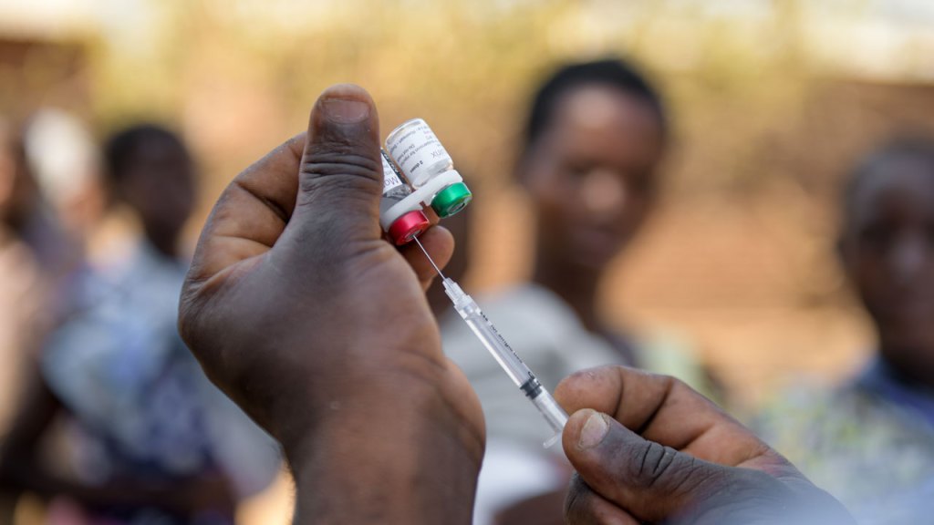 A close-up of hands holding two vaccine vials, one with a red cap and the other with a green cap, while drawing liquid into a syringe. The background is blurred with people standing.