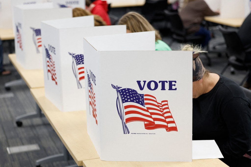 A row of white voting booths with the word "VOTE" and an American flag printed on the front. People are seated behind the booths, filling out ballots. The setting appears to be an indoor polling station with tables and chairs.
