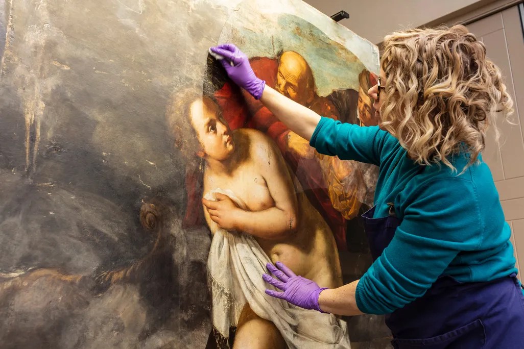 A woman wearing purple gloves and a teal sweater is carefully restoring a large classical painting depicting a partially nude figure draped in a white cloth, with other figures in the background. She is gently cleaning the surface of the artwork.