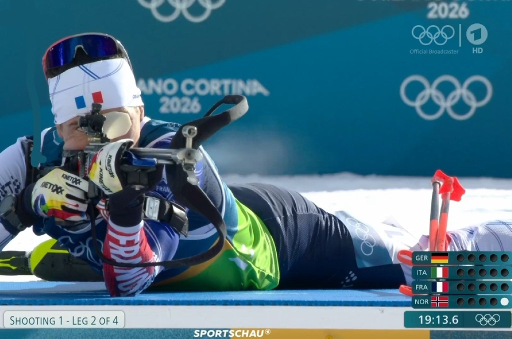 A biathlete in a prone shooting position aiming a rifle, wearing a white headband with the French flag and blue, green, and red sportswear. The background shows "MILANO CORTINA 2026" and Olympic rings. A scoreboard on the right displays team standings for Germany, Italy, France, and Norway, with Norway having one target hit. The timer reads 19:13.6, and the event is labeled "SHOOTING 1 - LEG 2 OF 4.
